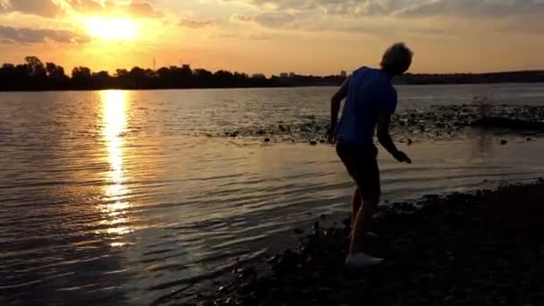 Sportive Man Stands on a Riverbank at Sunset. il jette une pierre — Video