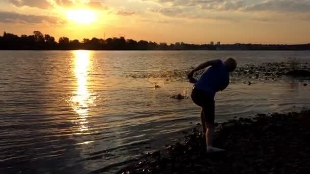 Un hombre inteligente se para en una orilla del río al atardecer. arroja una piedra — Vídeos de Stock