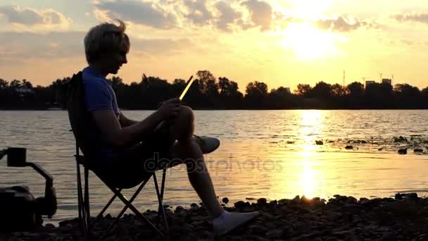 Heldere Man zit op een stoel vouwen en kijkt naar zijn Tablet pc bij zonsondergang — Stockvideo