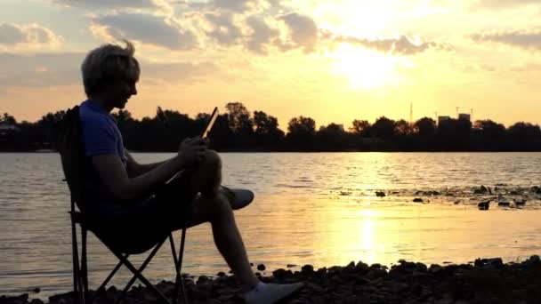 Έξυπνος άνθρωπος κάθεται σε μια καρέκλα αναδίπλωσης και εξετάζει τον υπολογιστή Tablet στο ηλιοβασίλεμα — Αρχείο Βίντεο