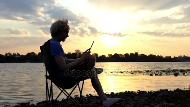 Ξανθός άντρας κάθεται σε μια καρέκλα αναδίπλωσης και εξετάζει τον υπολογιστή Tablet στο ηλιοβασίλεμα — Αρχείο Βίντεο