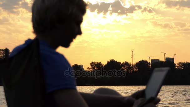 Man zit op een opvouwbare stoel en kijkt naar zijn Tablet pc bij zonsondergang onscherp — Stockvideo