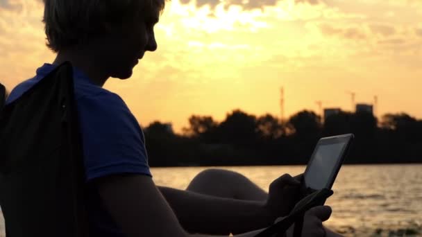 Письменник сидить на стільці розкладні і дивиться на його планшетного ПК на заході сонця — стокове відео