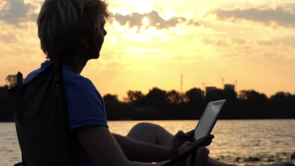 El hombre brillante se sienta en una silla plegable y mira su PC Tablet al atardecer — Vídeo de stock