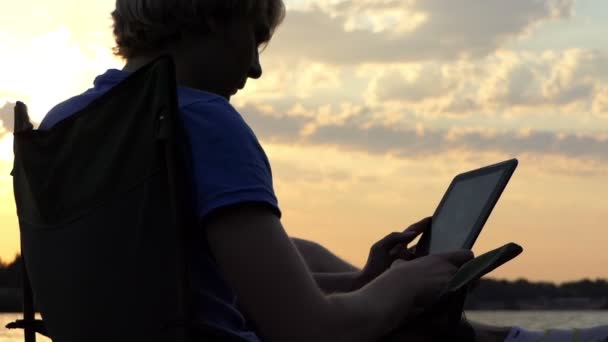 Junger Blogger sitzt auf einem Stuhl und blickt auf sein Tablet bei Sonnenuntergang im Slo-mo — Stockvideo