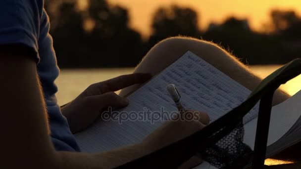 Giovane uomo si siede su una sedia pieghevole e scrive in un copybook al tramonto — Video Stock