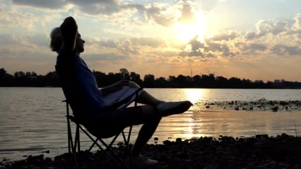 Giovane uomo si siede, legge e graffia il suo Nape su una riva del fiume al tramonto — Video Stock