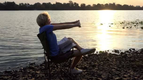 Jeune homme fait un exercice relaxant dans une chaise pliante au coucher du soleil — Video