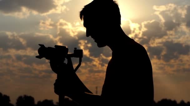 Cameraman jovem está em Perfil, Tunes Camera, e sorrisos ao pôr do sol — Vídeo de Stock