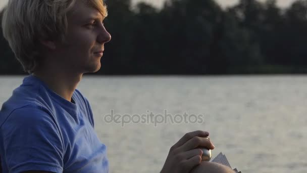 Man dricker kaffe och drömmar i en hopfällbar stol med hans anteckningar i solnedgången — Stockvideo