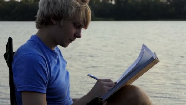Hombre rubio se sienta, lee y escribe sus notas en una orilla del río al atardecer en Slo-Mo — Vídeos de Stock