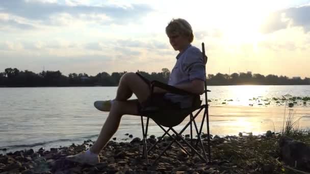 Joven se sienta en una silla y escribe su obra maestra en una orilla del río — Vídeo de stock