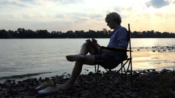 Blonder Mann sitzt an einem schönen Flussufer und schreibt in seinen Notizblock — Stockvideo
