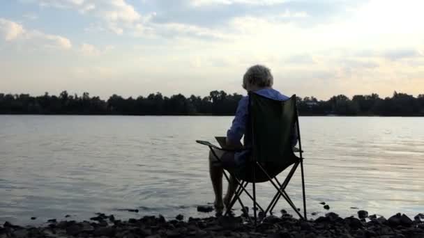 El hombre creativo se sienta, lee y escribe sus notas en una orilla del río al atardecer — Vídeos de Stock