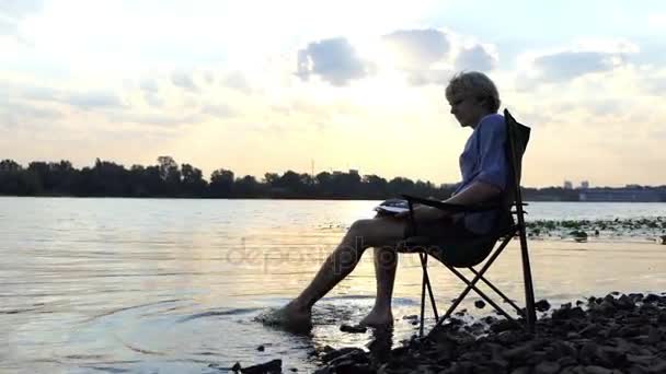 Φωτεινό άνθρωπος κάθεται και γράφει κάτω του Σημειωματάριου (Notepad) σε ένα ωραίο Riverbank — Αρχείο Βίντεο