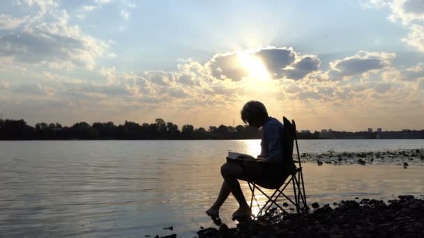 Escritor se sienta, lee y pone sus notas en una orilla del río al atardecer en Slo-Mo — Vídeo de stock