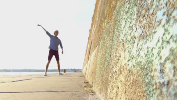 Junger Mann hebt die Hände und tanzt auf einem Flussufer mit einer hohen Mauer im Slo-mo — Stockvideo