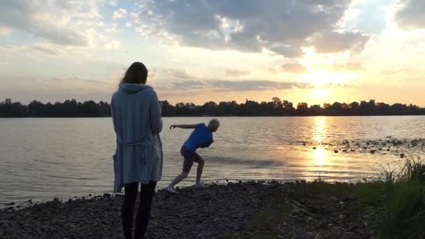 Jonge vrouw en Man staan op een rivieroever. Hij gooit een steen — Stockvideo
