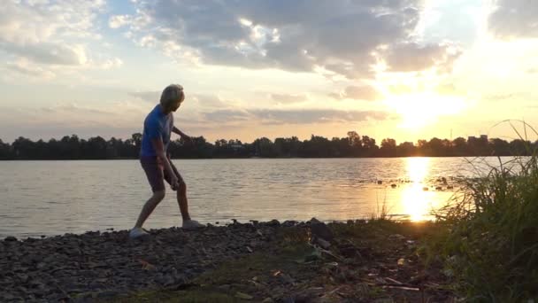 Молодой человек стоит босиком в воде на берегу реки и бросает камень в Сло-Мо — стоковое видео