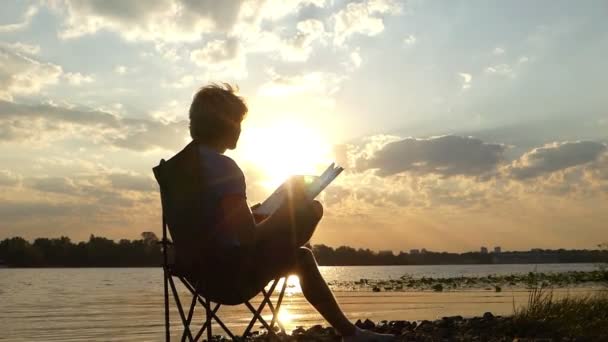 Man zit, denkt, schrijft iets in zijn Kladblok, op een rivieroever in Slo-Mo — Stockvideo