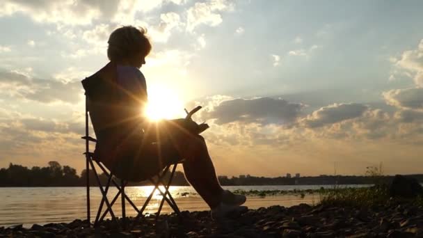 Jongeman zit, denkt, leest iets in zijn Kladblok, op een rivieroever in Slo-Mo — Stockvideo