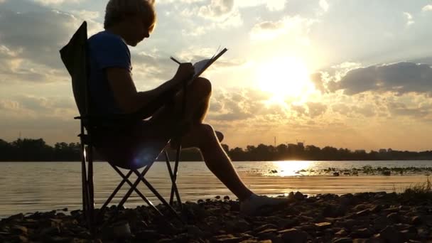 Ung Man sitter på en hopfällbar stol på floden, skriver vid solnedgången i Slo-Mo — Stockvideo