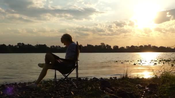 Bright Man senta-se em uma cadeira dobrável na margem do rio, escreve ao pôr do sol — Vídeo de Stock