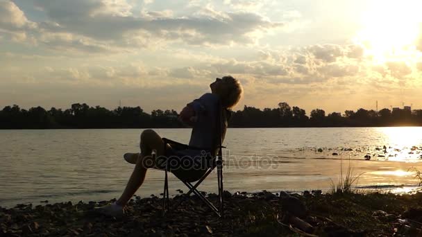 Молодой человек сидит и растягивается на берегу реки на Сансет в медленном движении — стоковое видео