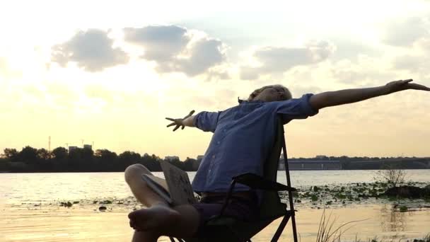 Homem alegre se senta e se estica em uma margem do rio em um pôr do sol esplêndido em Slo-Mo — Vídeo de Stock