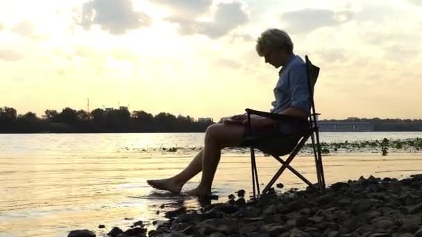 Νεαρός άνδρας κάθεται σε ένα Riverbank, γράφει, και κρατά το πόδι του στο νερό — Αρχείο Βίντεο