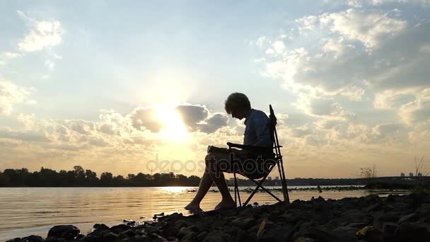 El hombre inteligente se sienta en una orilla del río, escribe, y mantiene su pie en el agua en Slo-Mo — Vídeo de stock