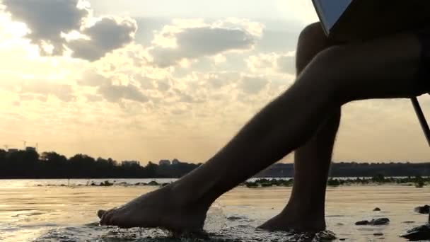 Jongeman zit op een rivieroever, schrijft, en beweegt zijn voet in het Water in Slo-Mo — Stockvideo