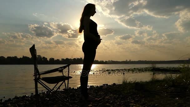 Joven embarazada golpea su vientre, se para en una orilla del río al atardecer en Slo-Mo — Vídeo de stock
