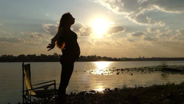 Junge schwangere Frau steht und streckt sich am Ufer des Flusses bei Sonnenuntergang in slo-mo — Stockvideo