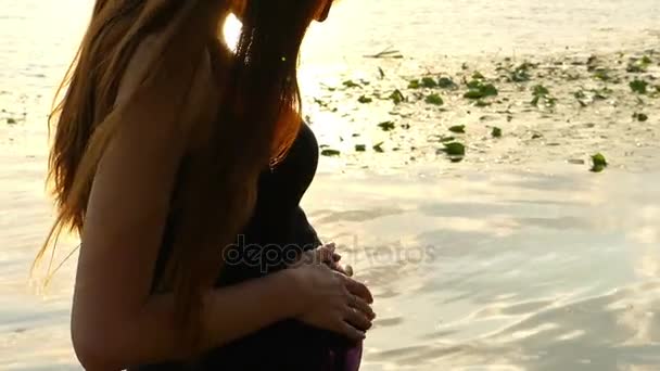 Mulher grávida bonita bate sua barriga, fica em uma margem do rio ao pôr do sol — Vídeo de Stock