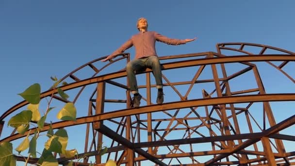 Joven sentado en una construcción metálica al atardecer en slo-mo — Vídeo de stock