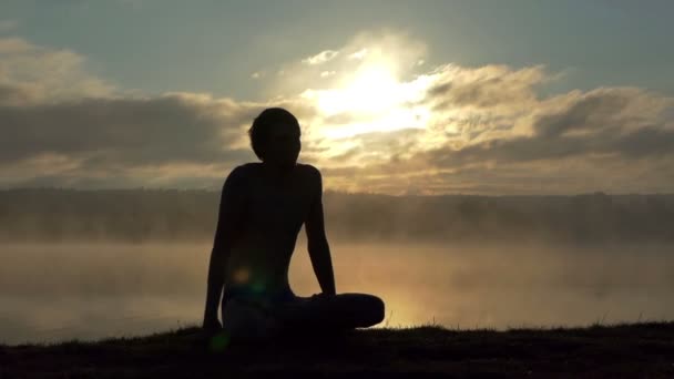 Joven se sienta en una orilla del lago y practica yoga al atardecer en cámara lenta — Vídeo de stock