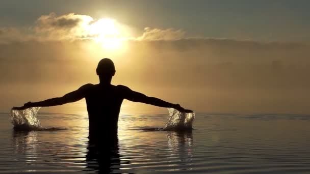 Vrolijke man werpt water omhoog in een meer bij zonsondergang in slo-mo — Stockvideo