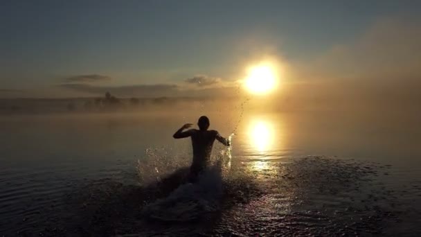 Щасливий чоловік бігає і стрибає в озері на заході сонця в шлю-мо — стокове відео