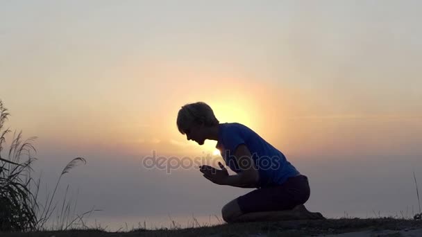 Jovem ajoelha-se e reza em um banco do lago ao pôr do sol em slo-mo — Vídeo de Stock