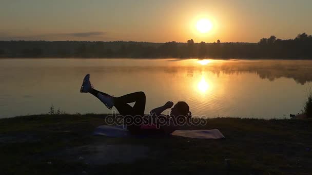 Chica entrena sus abdominales al atardecer cerca del lago . — Vídeos de Stock