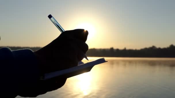 Female hand writes down something in a pad at sunset in 4k — Stock Video