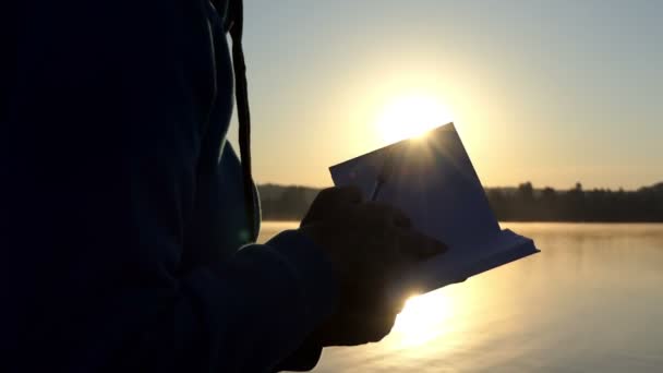 La mano femenina abre un bloc de notas en un lago al atardecer en 4k — Vídeo de stock