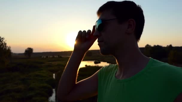 Молодий чоловік на заході сонця знімає сонцезахисні окуляри у повільному русі . — стокове відео