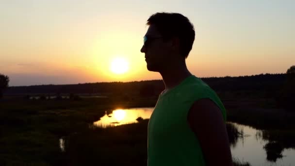 Homme en lunettes de soleil jouit d'un beau coucher de soleil près du lac . — Video