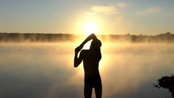 Young man stretches out his arms on a lake bank at sunset — Stock Video