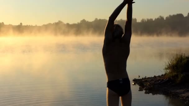 Joven hombre calienta sus brazos en una orilla del lago al atardecer — Vídeos de Stock