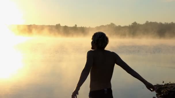 Молодий чоловік піднімає руки на березі озера на заході сонця — стокове відео