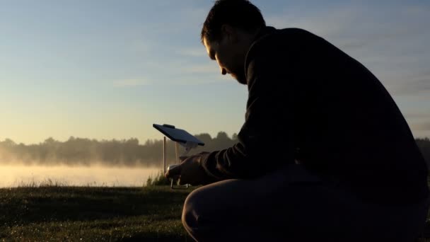 Joven se sienta y mira un tablero de drones en una orilla del lago — Vídeo de stock