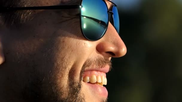 Joven feliz en gafas de sol mira hacia adelante y sonríe — Vídeos de Stock